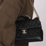 Black quilted handbag with gold chain strap held by a person wearing a brown outfit.