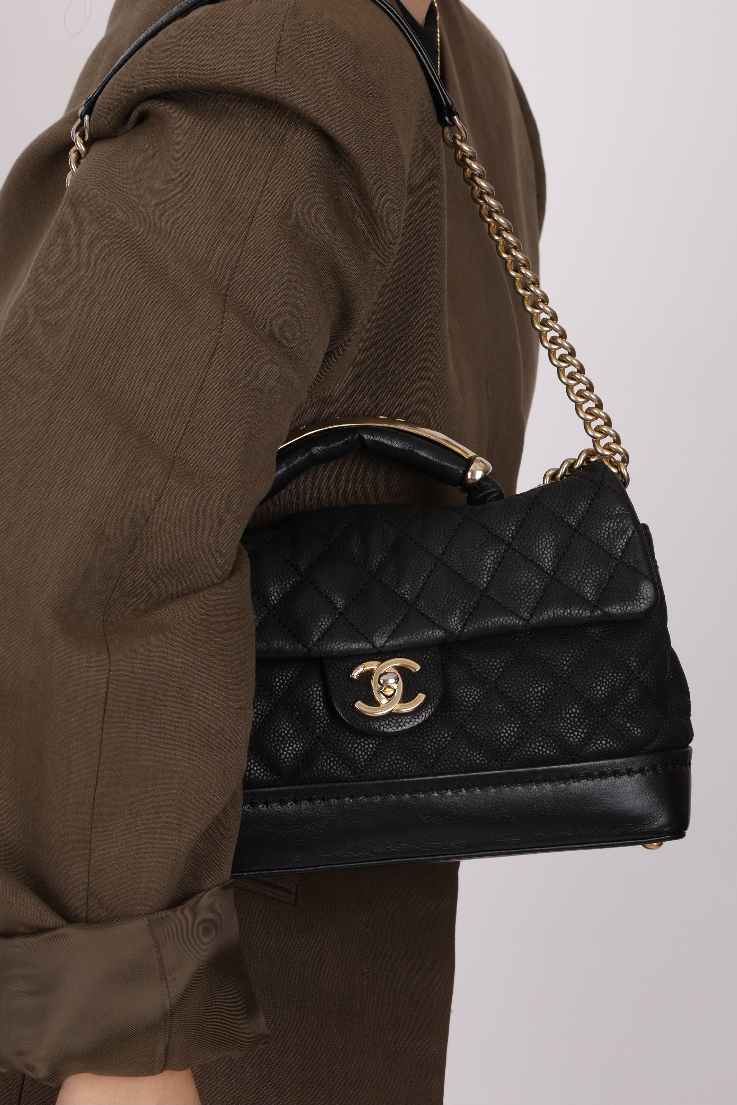 Black quilted handbag with gold chain strap held by a person wearing a brown outfit.
