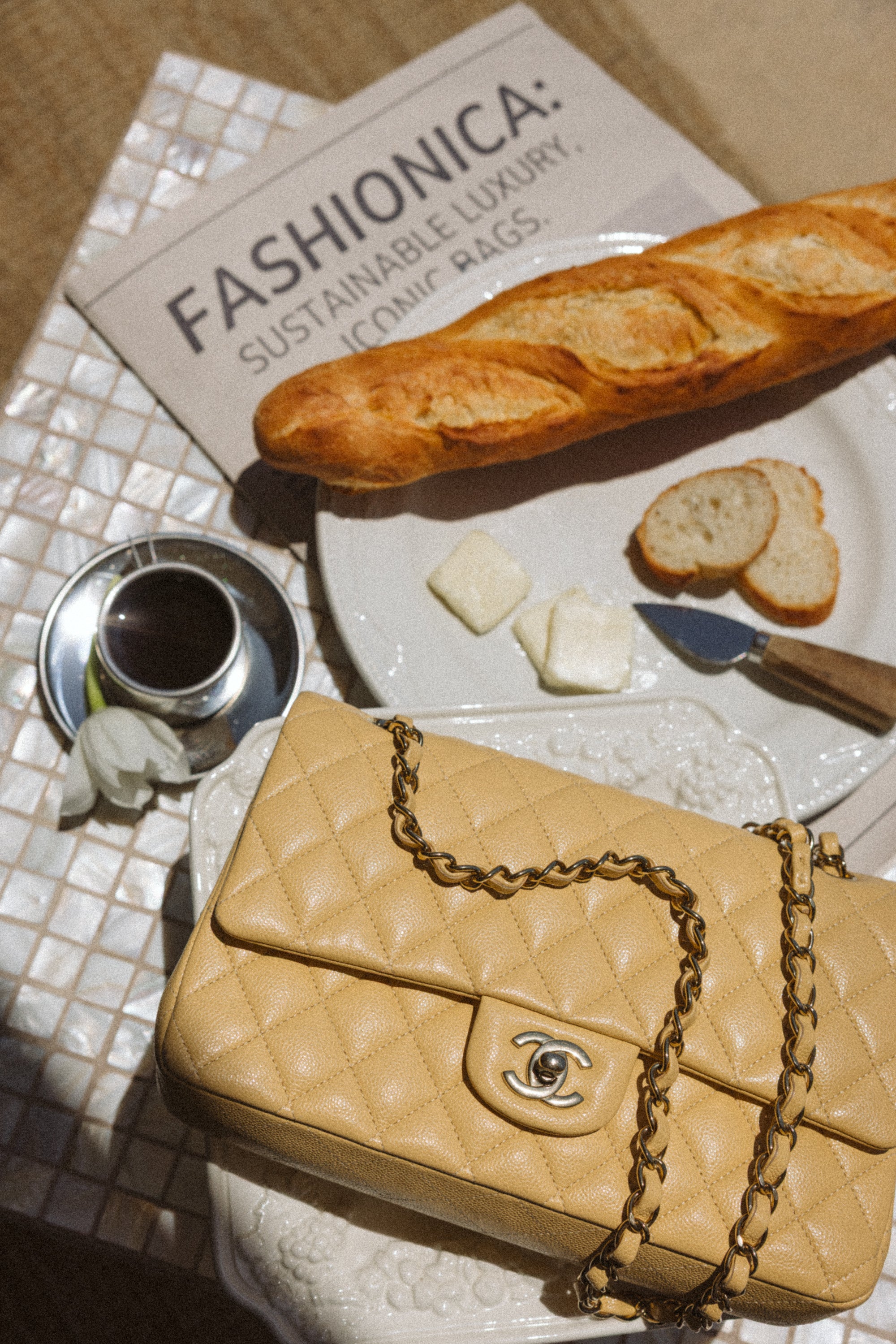 Beige handbag with gold chain on a table with a newspaper, baguette, and coffee.